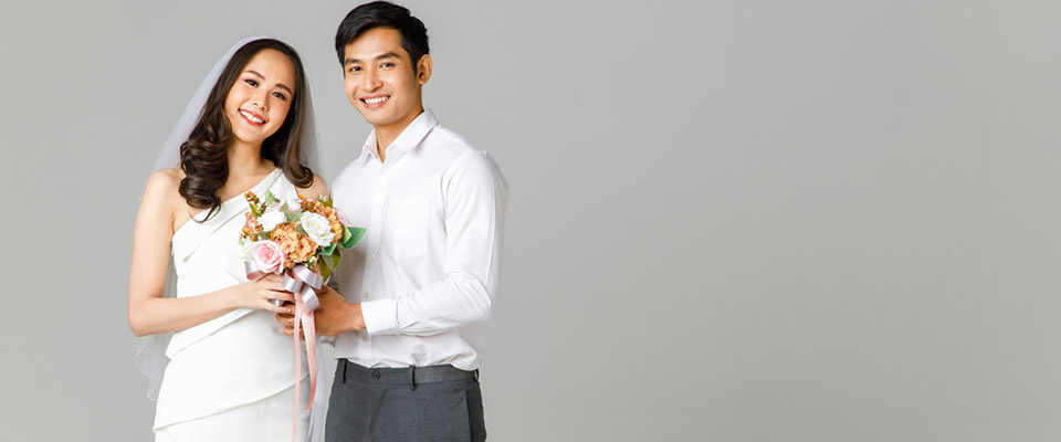 Young attractive Asian couple, man wearing white shirt, woman wearing white dress with wedding veil holding bouquet of flowers together. Concept for pre wedding photography.